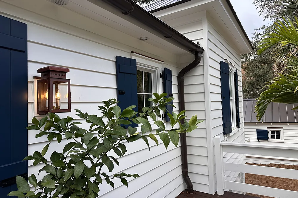 White coastal-style house exterior with horizontal white siding, navy blue shutters, black gutters, and a bronze lantern-style wall sconce mounted beside a window, surrounded by greenery and palm trees.