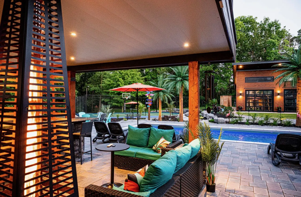 Covered outdoor patio with teal and green cushioned wicker seating, a fire feature, and a view of a backyard pool, sport court, and pool house at dusk.