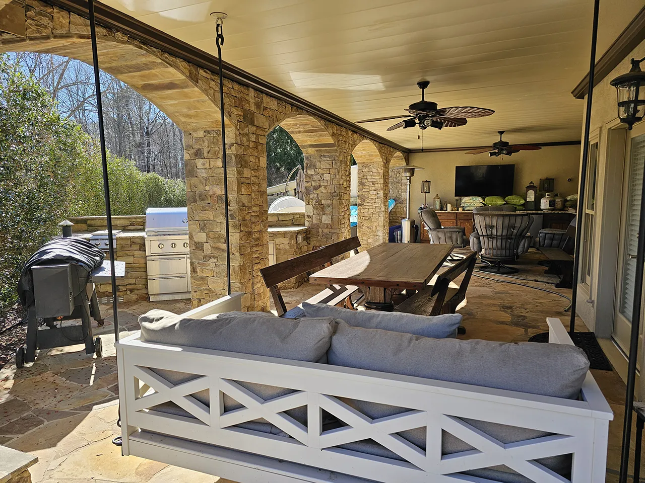 Underdeck patio with ceiling fans, outdoor sofa, and canvas curtain panels beneath a second-story wood deck with stairs leading down to the yard.