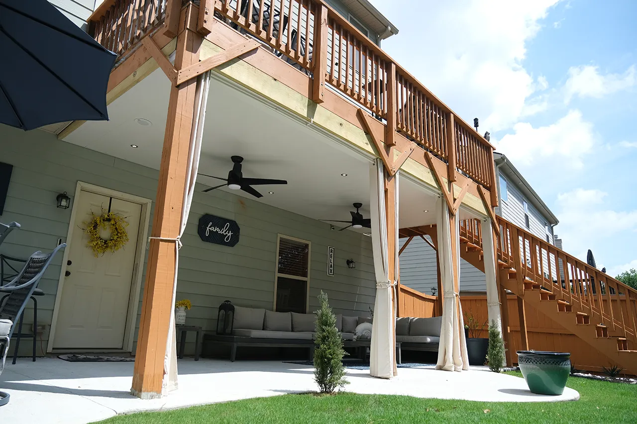 Covered stone-arched patio with an outdoor kitchen, dining table, and lounge seating overlooking a wooded backyard.
